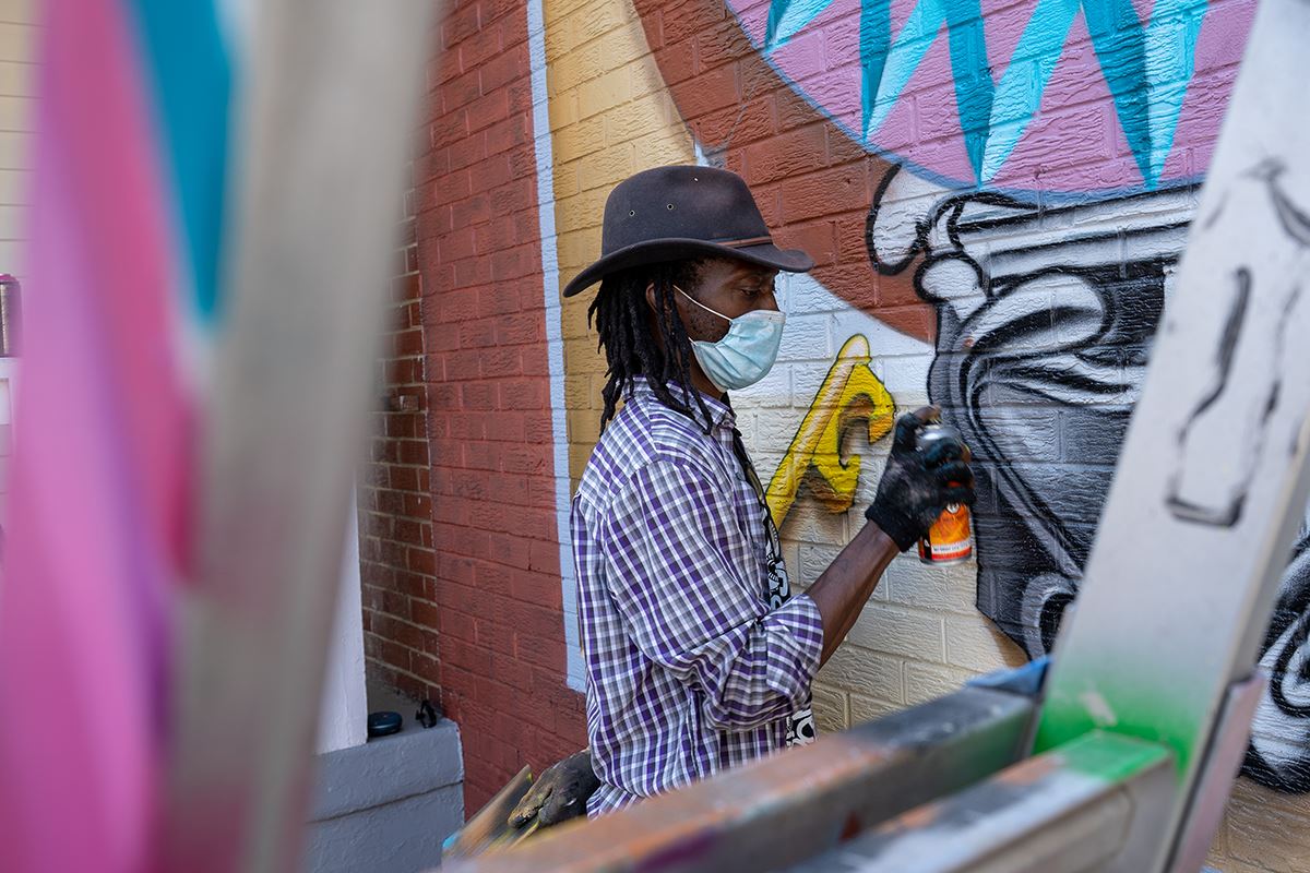 Artist Eric B. Ricks, wearing a fedora and a protective mask spray paints details of a large mural.